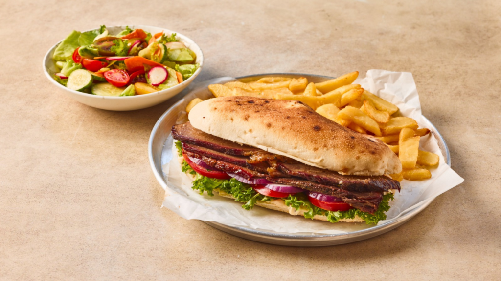 כריך בשר פרוס (כמו בריסקט או אסאדו) בלחמנייה פריכה עם חסה, עגבניה ובצל סגול, מוגש על מגש מתכת לצד צ'יפס זהוב וקערת סלט ירקות טרי